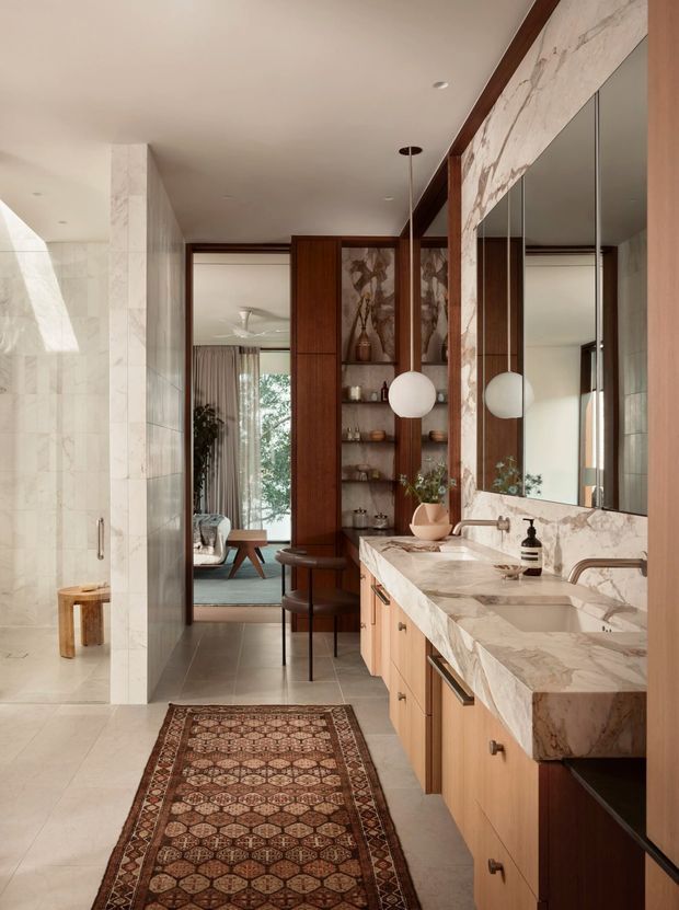A refined, spacious bathroom with a long double vanity in veined marble, light timber cabinetry, large mirrored panels, marble wall cladding, and spherical pendant lights. A freestanding bathtub is partially visible on the left, while a timber-and-marble screen opens toward a sitting area beyond. The scheme combines warm timber, stone, bronze-toned details, and a patterned runner for a luxurious contemporary interior.