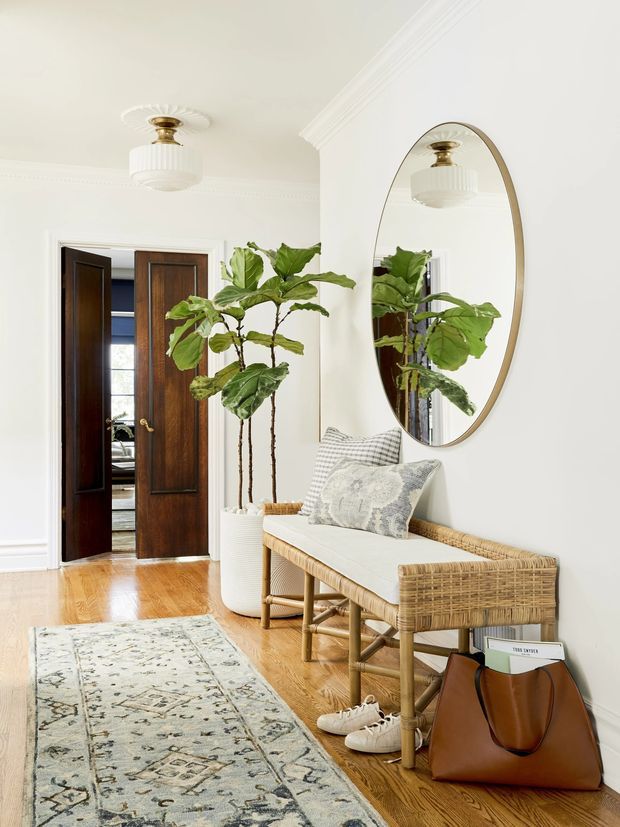 A bright, refined residential entry hall with polished timber flooring, white walls and decorative ceiling moulding. A rattan bench with a pale cushion sits beneath a large oval framed mirror, accompanied by a tall potted fiddle-leaf fig. Dark timber double doors open toward another room, while a patterned runner, woven textures and a leather tote add warmth. A vintage-style pendant ceiling light and brass-toned details complete the space.