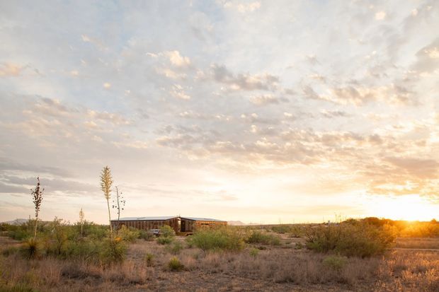 Wide landscape view of a low, elongated single-storey building set in a dry, sparsely vegetated rural landscape. The structure has a simple horizontal profile with what appears to be a low-pitched or flat roof and dark timber-toned exterior elements. The image is dominated by an expansive sunset sky and warm low sunlight, with scrubby grasses and young trees surrounding the building.