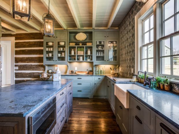 A spacious, warmly styled kitchen with a large central island, extensive grey-blue cabinetry, dark stone benchtops, open shelving and glass-front cupboards. The room features exposed timber ceiling beams, a rustic horizontal timber wall, patterned wallpaper, wide multi-pane windows, pendant lantern lighting, and dark timber flooring.