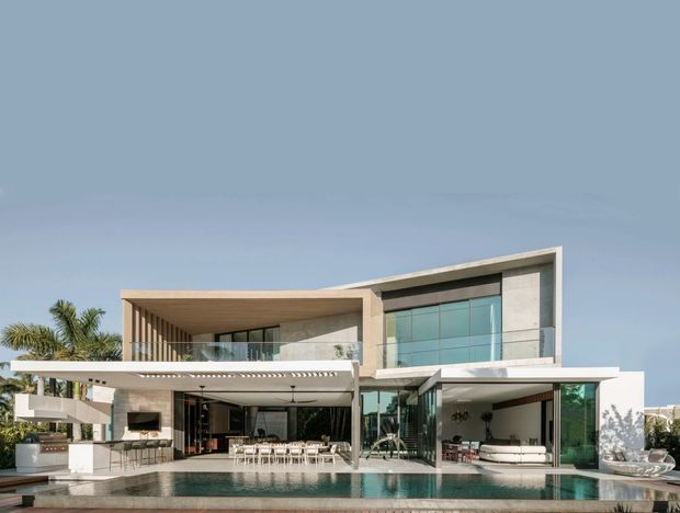 A contemporary multi-level luxury residence viewed from the pool terrace. The building has clean rectilinear volumes, extensive floor-to-ceiling glazing, a projecting flat-roofed entertaining canopy, pale rendered or concrete walls, timber-toned soffits, and a large swimming pool in the foreground. Palm landscaping and outdoor dining and lounge areas are visible around the open-plan ground floor.