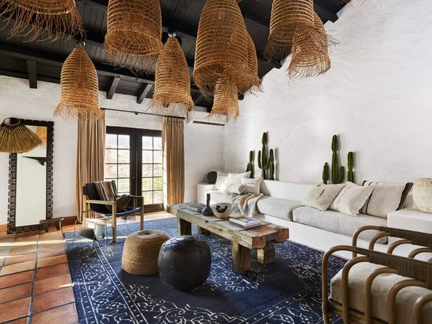 A spacious, eclectic living room with white textured walls, exposed dark ceiling beams, terracotta tile flooring, and a large dark patterned rug. A long light-coloured sofa, rustic timber coffee table, leather ottomans, woven seating, tall cacti, and numerous oversized woven pendant lights create a warm desert-inspired interior.