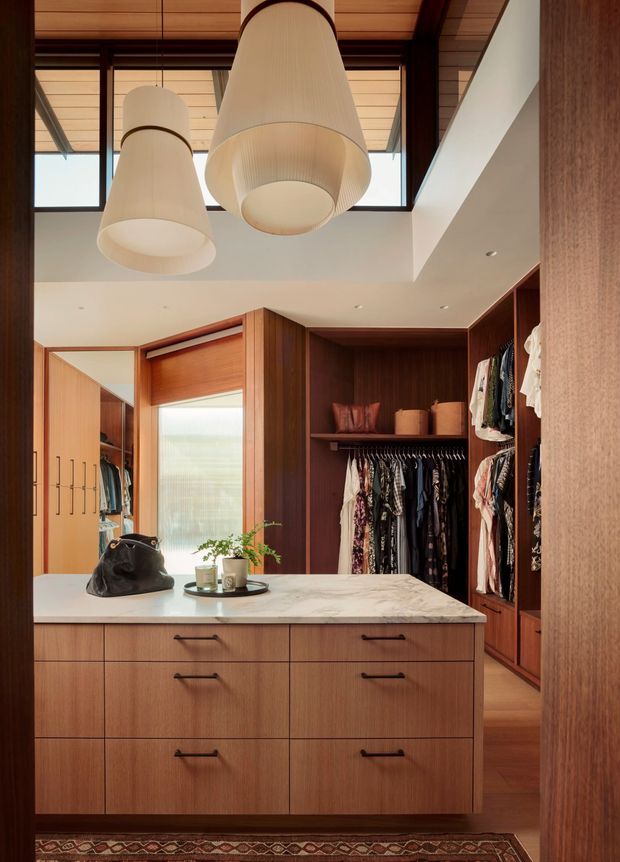 A refined walk-in wardrobe with warm timber cabinetry, open hanging rails, drawers, and a large central island with a light stone benchtop. The room features a dramatic double-height ceiling, clerestory glazing, timber ceiling lining, and two oversized pendant lights, creating a warm, luxurious dressing space.