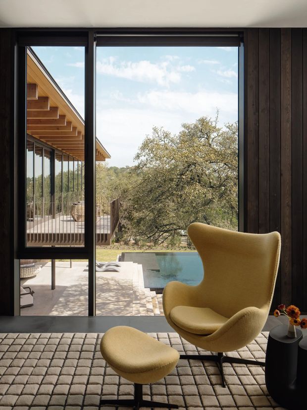 Interior view through expansive black-framed glazing toward a covered outdoor terrace and swimming pool, with a sculptural mustard lounge chair and ottoman in the foreground. The room features dark vertical timber wall panelling, a pale ceiling, and a large gridded neutral rug. The setting has a refined mid-century-inspired character with strong indoor-outdoor connection.
