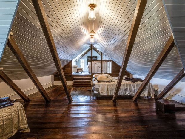 A rustic attic bedroom beneath a steeply pitched, white-painted vaulted ceiling. Exposed dark timber rafters frame polished dark timber flooring, with multiple beds, a small raised landing, and a distant sitting area visible. Warm pendant lights create a cozy, lodge-like atmosphere.