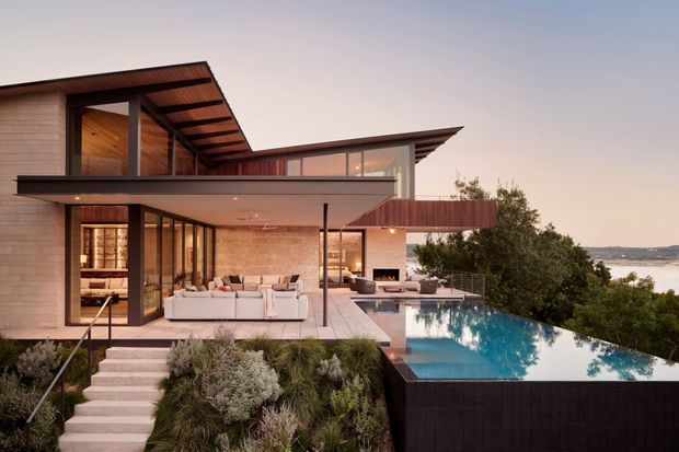 Contemporary multi-level waterfront residence with broad floor-to-ceiling glazing, warm timber cladding and soffits, pale masonry walls, and expansive cantilevered roof planes. The open living area is visible through the glazing, while a stepped landscaped garden leads to a dark-edged infinity pool overlooking the water.