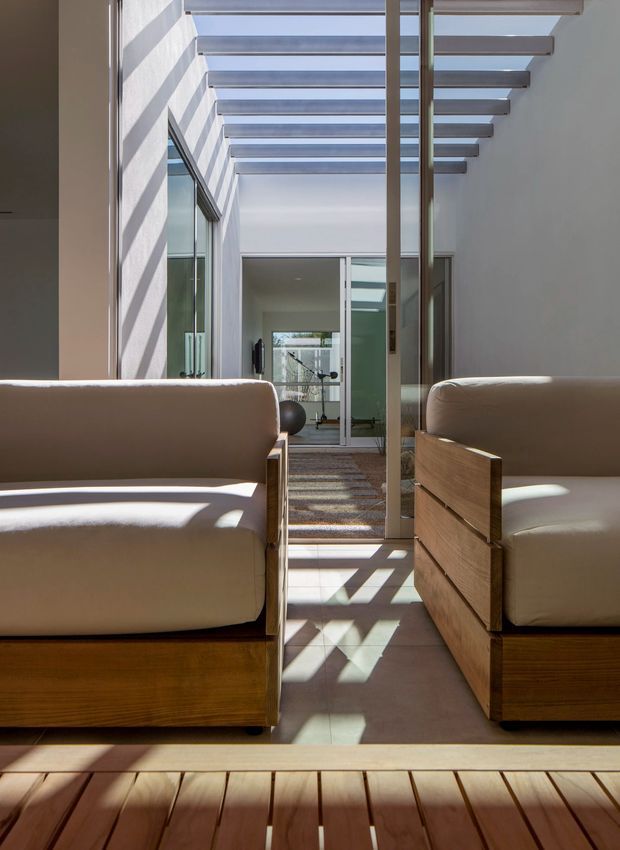 Bright modern interior corridor or living space framed by large glazing and a linear pergola-style roof structure. Two substantial upholstered lounge chairs with warm timber bases sit in the foreground, while strong sunlight casts rhythmic geometric shadows across the pale tiled floor and timber threshold. Glass doors lead toward another minimalist room, creating a layered, airy composition.