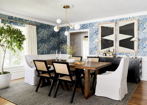 A bright, traditionally detailed dining room with a large rectangular dark timber dining table surrounded by upholstered slipcovered chairs and black-framed woven-back chairs. Blue botanical wallpaper covers the walls above white decorative wainscoting, with white curtains, timber flooring, a dark sideboard, graphic framed artwork, and a sculptural globe pendant light. A large potted indoor tree sits beside the window.