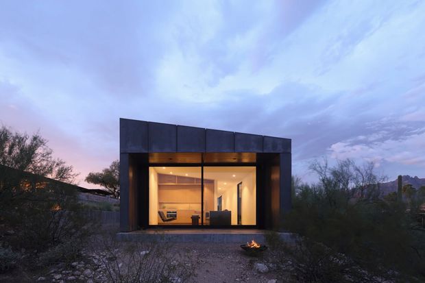 A compact modern pavilion-like building photographed at dusk, set within a sparse desert landscape. The dark monolithic facade has a slightly angled flat roof and a broad floor-to-ceiling glazed opening, revealing a warmly lit minimalist interior. A small outdoor fire sits in the gravel foreground, with native scrub surrounding the structure.