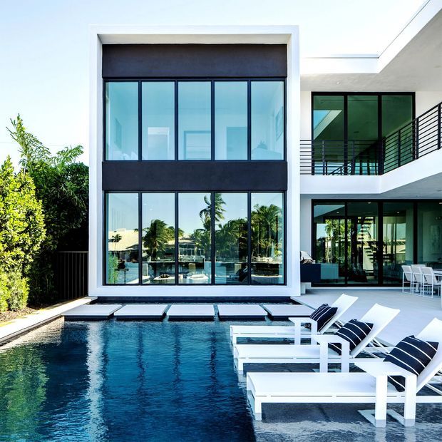 A bright contemporary multi-storey residence viewed from the pool courtyard. The facade features a tall white-framed glazed volume with dark horizontal bands, extensive black-framed windows, and an upper-level balcony with black metal railings. A rectangular swimming pool occupies the foreground, bordered by pale paving and white pool loungers with dark striped cushions. The setting includes lush planting and strong indoor-outdoor visual connections.