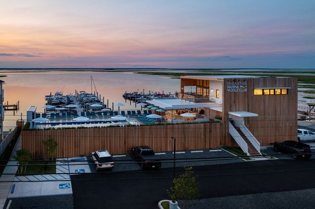 Aerial exterior view of a contemporary waterfront marina clubhouse or commercial building at dusk. The building has a warm timber-clad facade, a flat roof, large glazed openings, rooftop or elevated outdoor seating, and an external stair. Boats, pontoons, parking, and expansive tidal water are visible around the building.