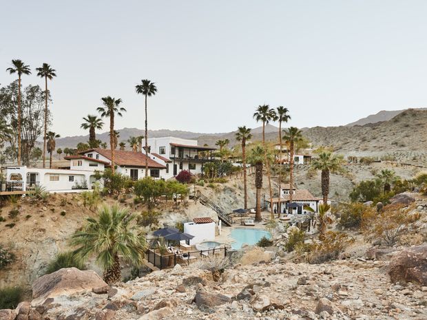 A hillside residential property set in a dry, rocky desert landscape, with several low-rise white buildings, red-tiled roofs, tall palm trees, landscaped terraces, and a small turquoise swimming pool. Rugged mountain terrain rises in the background.