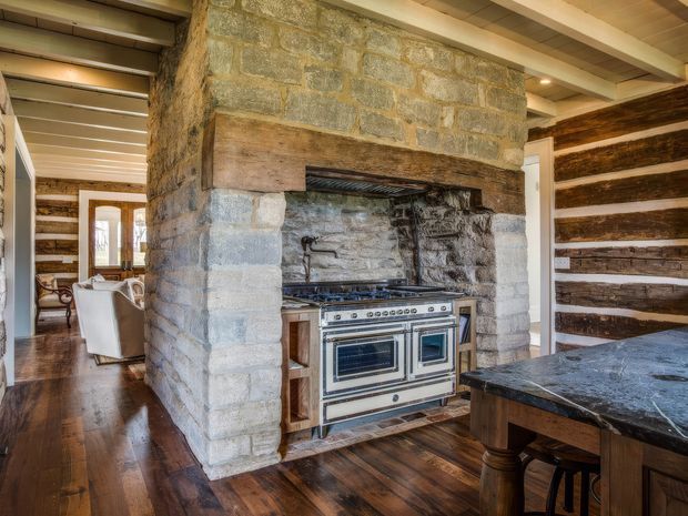 Rustic kitchen centered around a large exposed stone and timber masonry cooking hearth. A professional range sits beneath a dark stone backsplash and timber mantel, with a dark stone island in the foreground. The room features wide timber floorboards, exposed timber-and-plaster ceiling, and horizontal timber wall detailing. A dining or living area is visible beyond the kitchen.