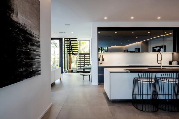 A bright, contemporary open-plan kitchen viewed from the adjacent living area. The kitchen features a large white island with a dark benchtop, integrated white cabinetry, a dark reflective splashback or mirrored wall panel, a brushed metal tap, and black wire bar stools. Recessed ceiling lights, large windows, and an open black-framed staircase create a clean, airy interior with strong black-and-white contrast.