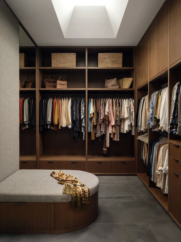 A sophisticated walk-in wardrobe with extensive dark timber built-in cabinetry, open shelving, drawers, hanging rails, and neatly arranged clothing and handbags. A large upholstered curved island bench occupies the foreground, while a roof skylight brings soft natural light into the space. The floor is finished in large dark grey tiles and the overall palette is warm, muted, and refined.