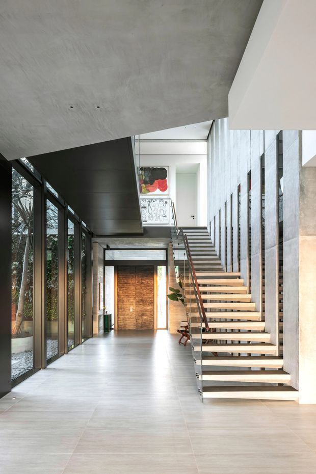 A spacious contemporary interior hallway and stairway with an open floating staircase, slim metal balustrade, extensive floor-to-ceiling glazing, pale tiled flooring, and exposed concrete ceiling. A timber entry door and upper-level artwork are visible beyond the stair. The space is bright, minimal, and architecturally focused, with strong vertical and horizontal lines.