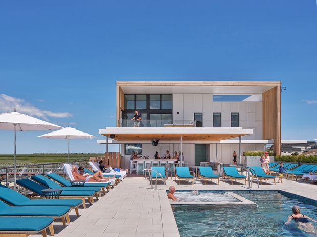 A bright contemporary multi-level poolside building with a rectilinear white facade, warm timber soffits and vertical timber screening. The image shows a large swimming pool, paved pool deck, turquoise lounge chairs, umbrellas, and people relaxing around the water under a clear blue sky.