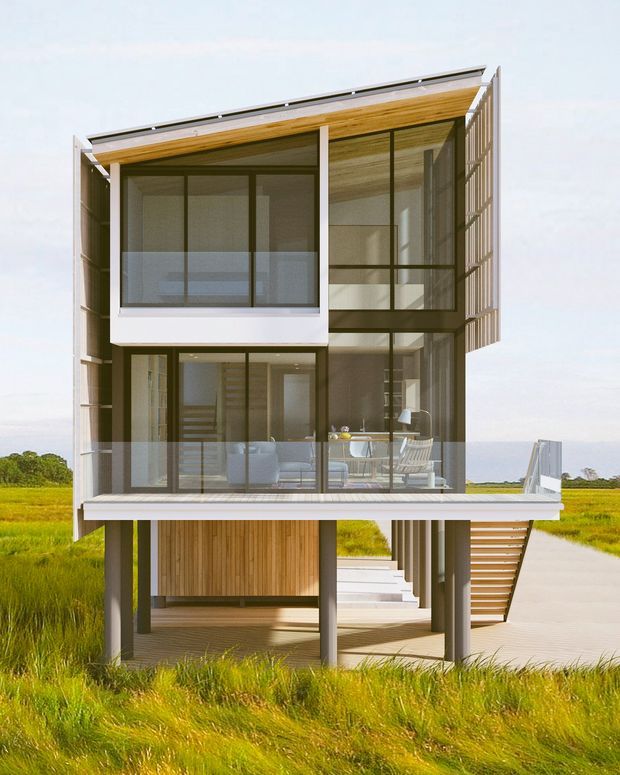A contemporary multi-level house elevated on slender posts in a grassy rural or coastal-looking landscape. The compact rectangular structure has extensive floor-to-ceiling glazing, dark window frames, timber-lined soffits and interior ceilings, timber cladding at the lower level, open decks with clear glass balustrades, and an external stair. The building presents a lightweight, transparent, rectilinear architectural form.