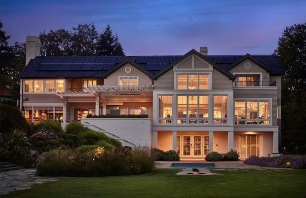 A large multi-storey suburban residence photographed at dusk, with a symmetrical gabled facade, dark metal pitched roof, grey horizontal cladding, extensive illuminated glazing, balconies and terraces, and a landscaped lawn and garden in the foreground. The house has a refined traditional exterior with contemporary-scale windows and detailing.