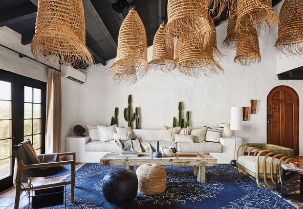 A styled living room with white textured plaster walls, dark exposed ceiling beams, tall black-framed glazed doors, a large white sectional sofa, patterned navy rug, rustic wooden coffee table, woven pendant lights, sculptural seating, cacti, and an arched timber door. The space blends relaxed southwestern, organic, and eclectic elements with strong natural textures and high visual contrast.