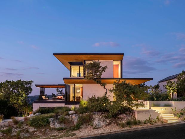 Twilight exterior view of a contemporary two-storey residence on a landscaped, rocky sloped site. The house features strong horizontal flat roof planes with deep timber soffits, warm interior lighting, large glazed openings, pale rendered walls, and an elevated terrace overlooking a distant landscape.