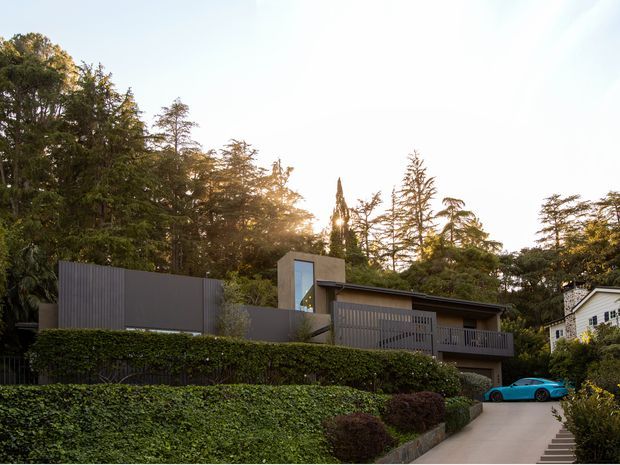 Exterior view of a contemporary multi-level residence set into a landscaped, wooded suburban site. The house features dark vertical cladding, a low-pitched roof, large glazing, a projecting upper-level balcony, privacy screens, and a stepped driveway approach. Dense hedges and mature trees frame the property, with a neighbouring light-coloured house visible at right.