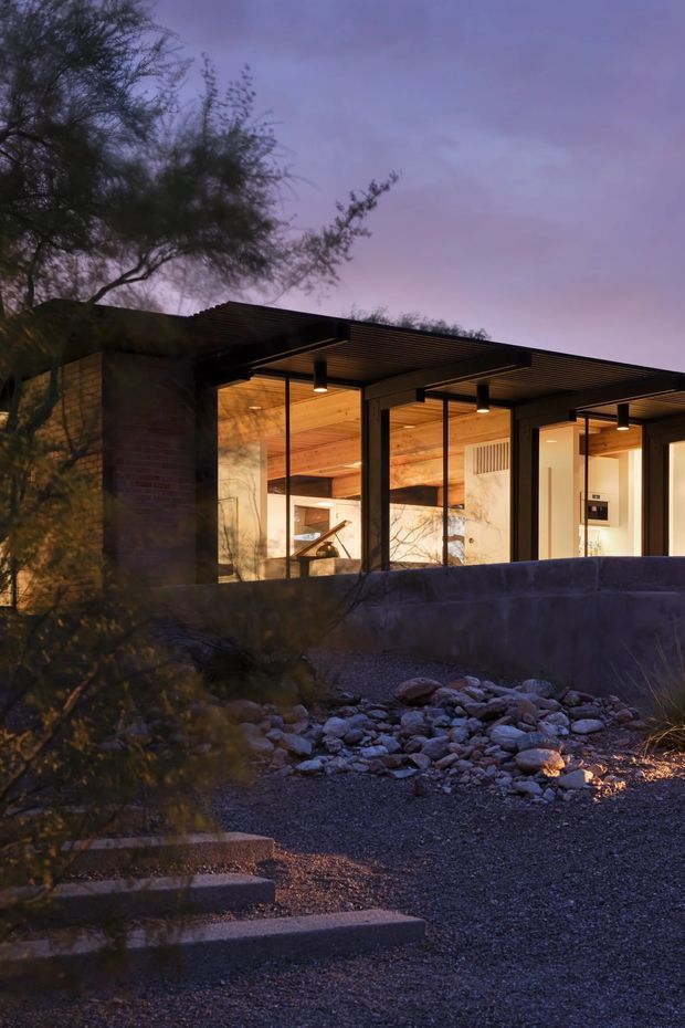 A dusk exterior view of a low-slung contemporary residence set into a rocky, landscaped site. The home has extensive floor-to-ceiling glazing, dark slender window frames, a broad flat projecting roof with a slatted underside, warm timber ceilings, exposed masonry walls, and softly lit interiors visible through the glass. Stone steps, gravel, rocks, and desert-like planting occupy the foreground beneath a purple evening sky.