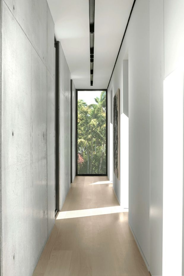 A narrow, minimalist interior hallway with pale timber flooring, smooth white walls, and a long recessed black linear ceiling light. Full-height glazing at the end opens views to lush tropical foliage, while framed artwork and concealed doors line the corridor. Strong sunlight creates bright geometric patches across the floor.