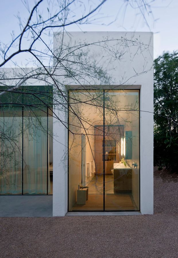 Exterior architectural photograph of a minimalist flat-roofed building at dusk, composed of smooth white rendered walls and large floor-to-ceiling glazed openings with slim dark frames. The warmly lit interior is visible through the glazing, including a bathroom vanity, mirrors, towels, shelving and timber flooring. Bare branching trees partially obscure the facade, while gravel landscaping surrounds the building.