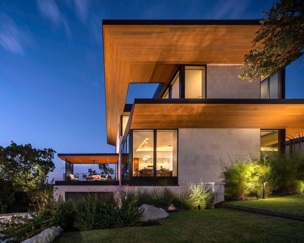 Twilight exterior view of a contemporary multi-level residence with dramatic cantilevered roof planes and broad timber-lined soffits. The facade combines pale masonry walls, dark window frames, extensive glazing, and sheltered outdoor living terraces. Low landscaped planting and a stepped lawn surround the house, with warm interior and exterior lighting creating a refined evening atmosphere.