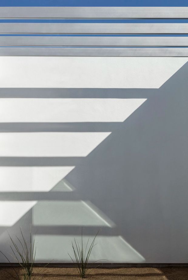 Abstract architectural detail showing a pale grey-white wall or facade with strong horizontal and diagonal bands of sunlight and shadow. Narrow blue-grey horizontal trim lines appear near the top, while sparse ornamental grasses sit along a gravel or sandy base. The image is minimalist and graphic, with no clearly visible room interior.
