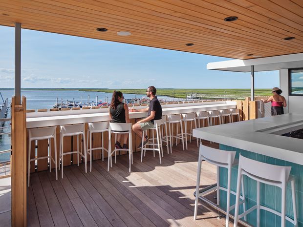 An open-air rooftop or waterfront hospitality terrace with a long bar counter and many white bar stools overlooking a marina and expansive coastal waterway. The terrace features dark timber decking, a warm timber slatted ceiling with recessed lights, slender dark structural posts, white built-in bar joinery, and a contemporary grey bar island with turquoise upholstered stools. The scene is bright and relaxed, with outdoor dining and panoramic water views.