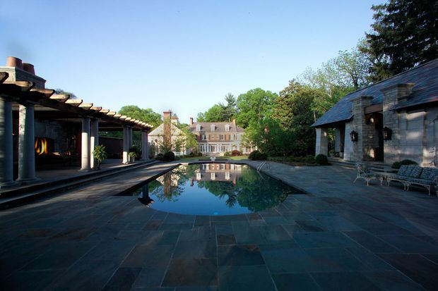 A formal landscaped courtyard centered on a long reflecting pool, framed by dark stone paving, a covered colonnade on the left, and a low slate-clad pavilion with outdoor seating on the right. A large traditional masonry house is visible beyond the pool, with mature trees surrounding the property. The composition has a refined, estate-like character and strong symmetrical axial planning.
