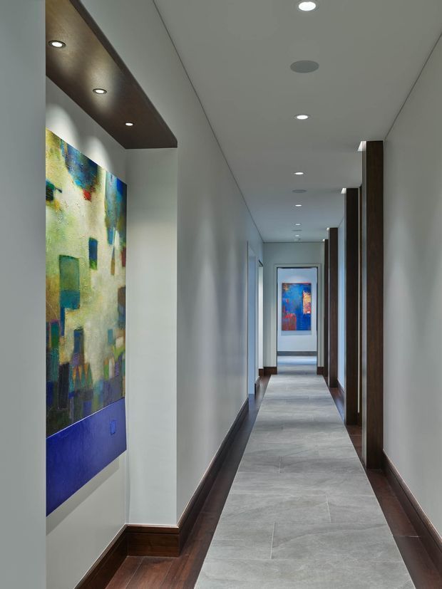 Long, narrow residential hallway with white walls, recessed ceiling lighting, dark timber skirting and door surrounds, and a pale grey tiled runner bordered by dark timber flooring. A large colourful abstract artwork is displayed beneath a dark timber ceiling pelmet with integrated spotlights, while additional artwork is visible at the far end.