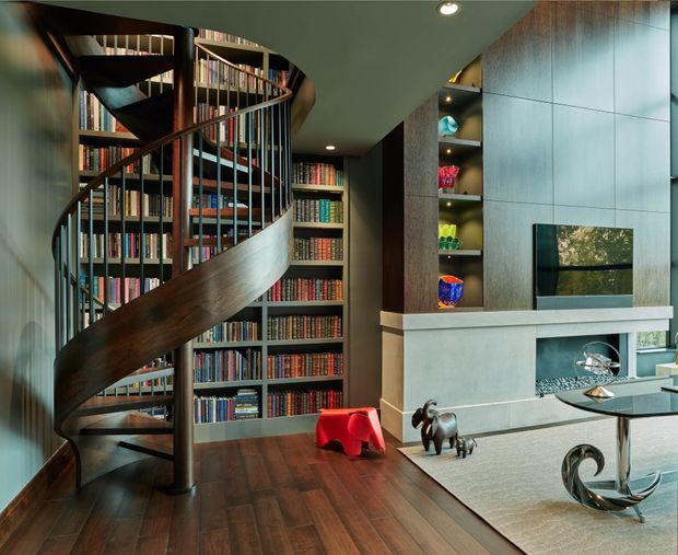 A sophisticated living room dominated by a sculptural dark timber spiral staircase winding through a floor-to-ceiling library. The room features dark wood flooring, extensive built-in bookcases, dark timber and grey wall cabinetry, a minimalist fireplace surround with integrated television, recessed display shelving, contemporary sculptural furniture, and a large pale rug. The composition has a refined, dramatic, high-contrast atmosphere with strong custom joinery and architectural detailing.