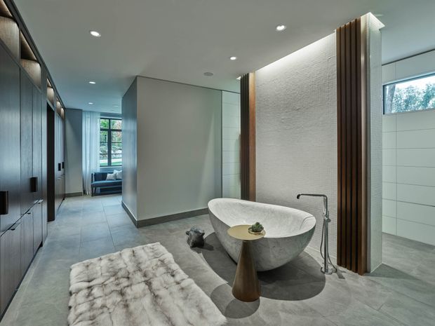 A refined contemporary bathroom featuring a freestanding sculptural bathtub, wall-mounted fixtures, large-format grey floor tiles, white tiled walls, and a vertical timber screen against a textured light wall. Dark built-in cabinetry lines the left side, while a corridor and seating area are visible beyond. Recessed ceiling lighting and large glazing create a calm, spacious atmosphere.
