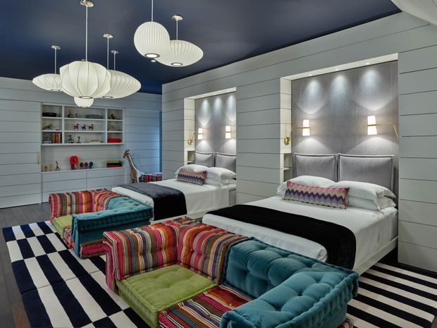 A bold, highly styled bedroom with two built-in beds set into illuminated wall niches. The room features white horizontal wall panelling, a dark navy ceiling, striped and vividly coloured upholstered seating at the foot of the beds, patterned cushions, and multiple sculptural pendant lights. A recessed open shelving unit displays books and small objects, while a graphic black-and-white striped rug covers the floor.