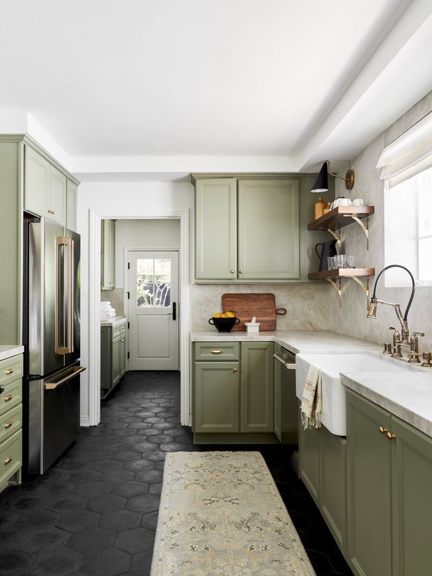 A refined galley-style kitchen with muted sage-green shaker cabinetry, dark hexagonal floor tiles, pale stone-look benchtops and backsplash, a farmhouse apron-front sink, stainless-steel refrigerator, brass hardware, open timber shelves, and a small adjoining utility or pantry area visible through the doorway. The room is bright and tailored, with traditional detailing and a warm, classic feel.