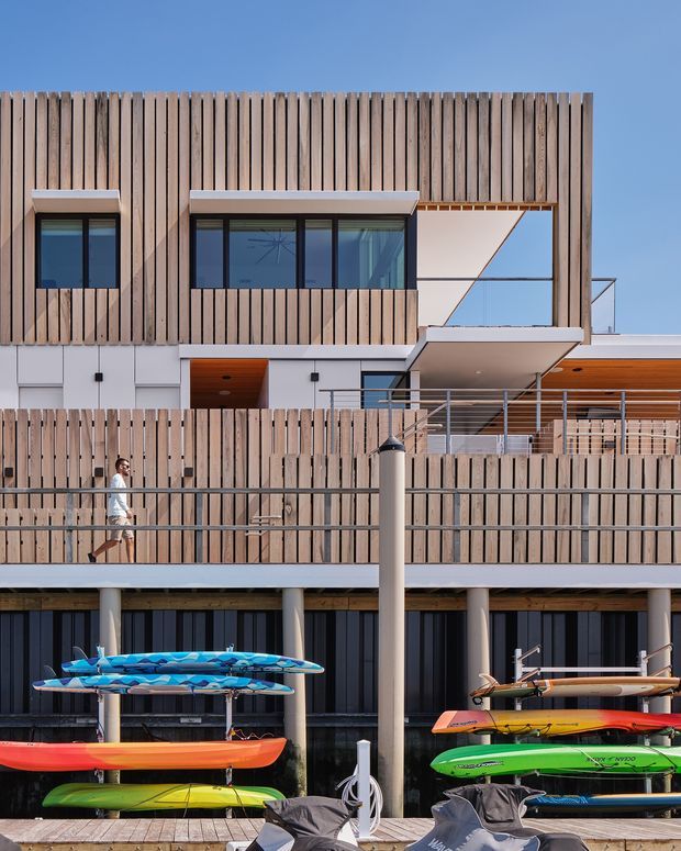 Exterior view of a contemporary multi-level building beside a waterfront or marina, featuring extensive vertical timber cladding, dark-framed glazing, white rendered walls, projecting balconies, metal balustrades, and a covered outdoor deck. Kayaks and paddleboards are stored in the foreground, with a person walking along the waterfront terrace. The composition emphasizes the building facade and its relationship to the waterside setting.