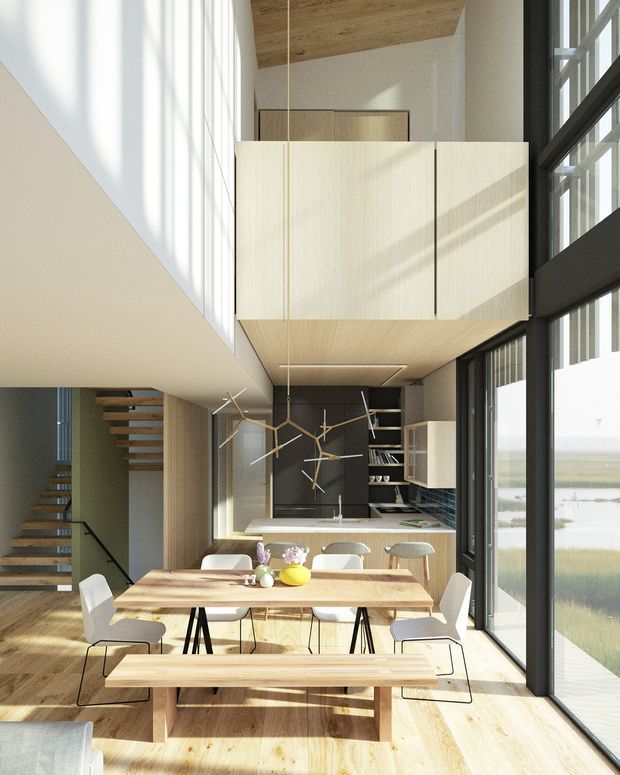 A bright, double-height contemporary open-plan interior combining a dining area and kitchen, with a floating mezzanine or upper-level enclosure above. The space features extensive black-framed floor-to-ceiling glazing, pale timber flooring and cabinetry, a timber ceiling, a large timber dining table with white chairs and bench seating, sculptural pendant lights, and a modern open-riser staircase. A calm waterside landscape is visible through the glazing.