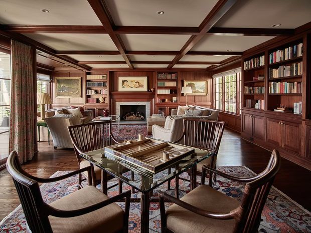 A richly appointed traditional library-living room with extensive dark timber wall panelling, built-in bookcases, a coffered timber-beam ceiling, and a central fireplace. A glass-topped dining or games table with wooden chairs occupies the foreground, while upholstered seating groups sit around the fireplace. Large multi-pane windows, patterned rugs, framed artwork, and classic lamps complete the warm, formal interior.