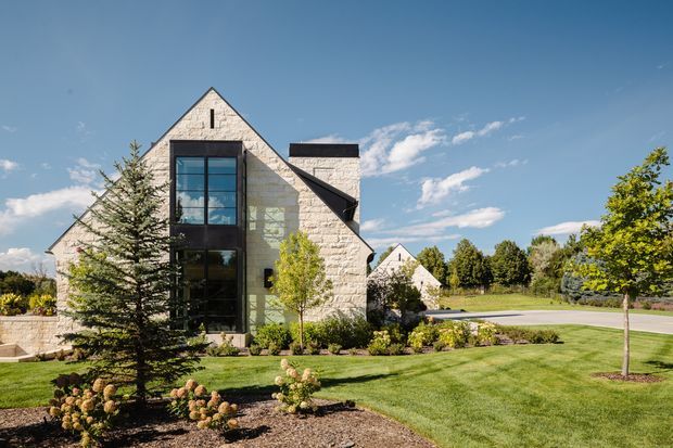 Exterior view of a substantial stone-clad house with steep gabled roof forms, a tall dark-framed glazed entry or stair volume, landscaped front lawn, mature planting, and a paved driveway under a clear blue sky.