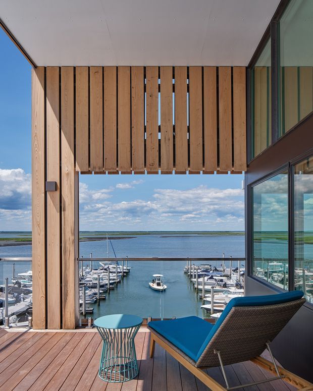 A modern waterfront balcony or terrace overlooking a marina, framed by warm vertical timber screening and large glazed openings. The space has timber decking, a blue outdoor lounge chair, and a small turquoise side table, with boats and calm water visible beyond.