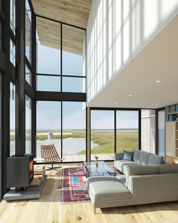 A bright, double-height contemporary living room with expansive black-framed floor-to-ceiling glazing overlooking a wetland landscape. The room features a pale timber ceiling and floor, white walls, a built-in black fireplace, grey sectional sofa and ottoman, a patterned area rug, glass coffee table, and an open timber shelving unit.