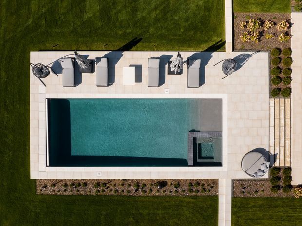 Aerial top-down view of a modern rectangular swimming pool surrounded by a pale paved terrace. Several loungers and outdoor chairs are arranged along the upper edge, with neatly landscaped garden beds, clipped lawn, and a straight pedestrian path framing the pool area.
