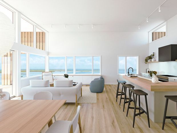 Bright open-plan coastal interior combining a kitchen, dining area and living room. The space features extensive floor-to-ceiling glazing with ocean views, pale timber flooring, white walls and cabinetry, a large timber kitchen island with seating, and minimalist contemporary furnishings. High ceilings and abundant natural light create an airy, relaxed atmosphere.