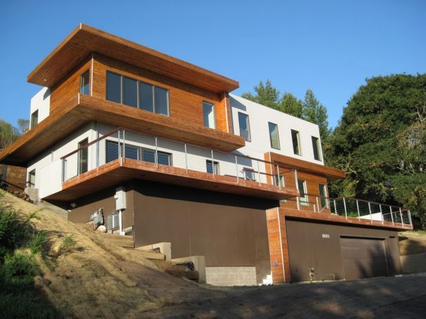 Exterior view of a contemporary multi-level hillside house with projecting timber-clad upper volumes, white rendered walls, broad balconies with metal balustrades, large glazed windows, and an integrated double garage. The home sits on a sloped site surrounded by mature vegetation under clear skies.