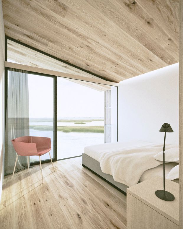 A bright, minimalist bedroom with pale timber flooring and matching timber-clad ceiling, white walls, and expansive dark-framed floor-to-ceiling glazing overlooking a calm coastal or wetland landscape. The room contains a low-profile upholstered bed with white bedding, a simple light timber bedside unit, a slim black floor lamp, and a muted pink accent chair. The design is restrained, warm, and strongly connected to the outdoor view.