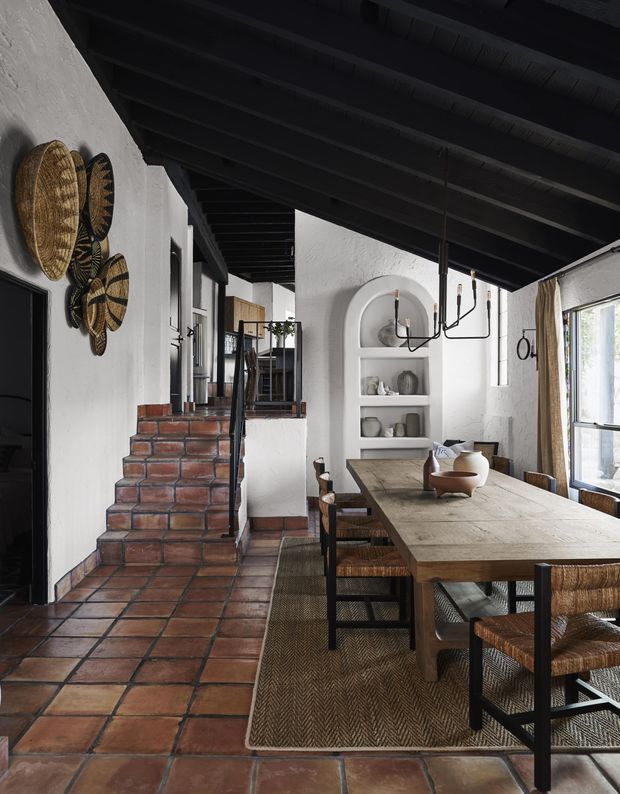 A warm, rustic dining area in a long open-plan interior. A large weathered timber dining table with woven-seat chairs sits on a natural fibre rug, beneath a black exposed-beam ceiling and a linear black chandelier. White textured plaster walls feature arched built-in shelving, while terracotta tile flooring continues through the space toward a short set of brick-edged steps. Woven wall baskets and ceramic objects add earthy character, with daylight entering through tall windows.