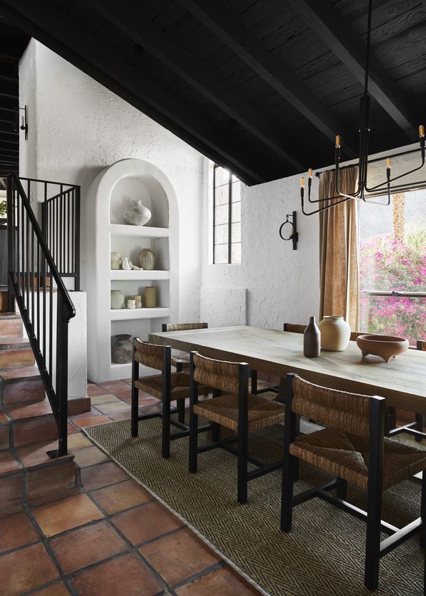 A warm, rustic dining area with a long rectangular timber dining table surrounded by woven-seat chairs. The room features terracotta tile flooring, heavily textured white plaster walls, a built-in arched display niche, black-painted exposed ceiling beams, and a black metal stair balustrade. Large windows with neutral curtains overlook flowering vegetation, while a linear sculptural chandelier hangs above the table.